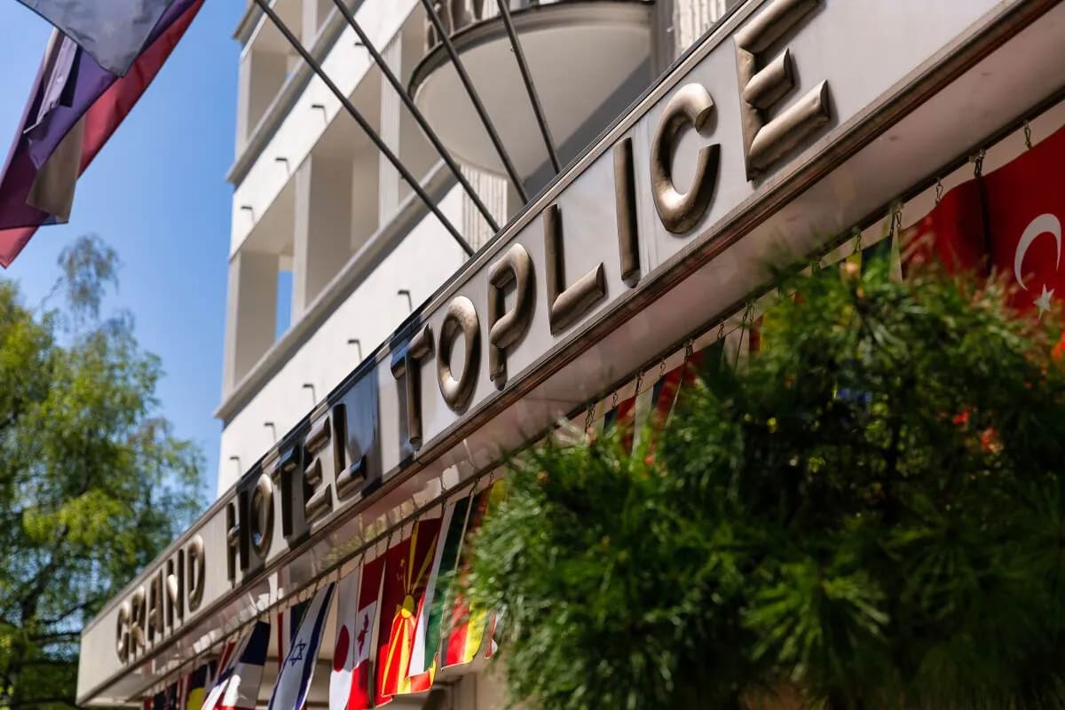 The sign of the Grand Hotel Toplice with several national flags hanging below, surrounded by greenery.