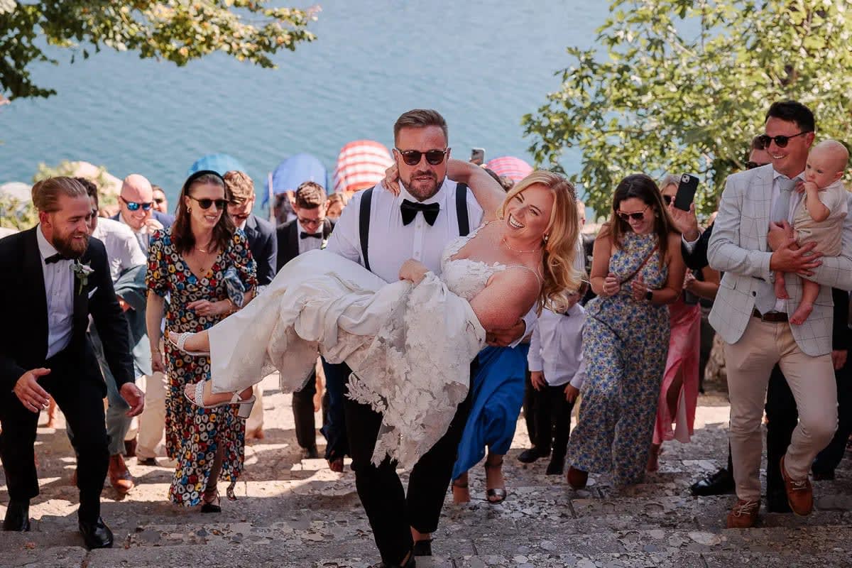 A groom carrying the bride up stone steps surrounded by wedding guests on Bled island with trees and pletna boats in the background.