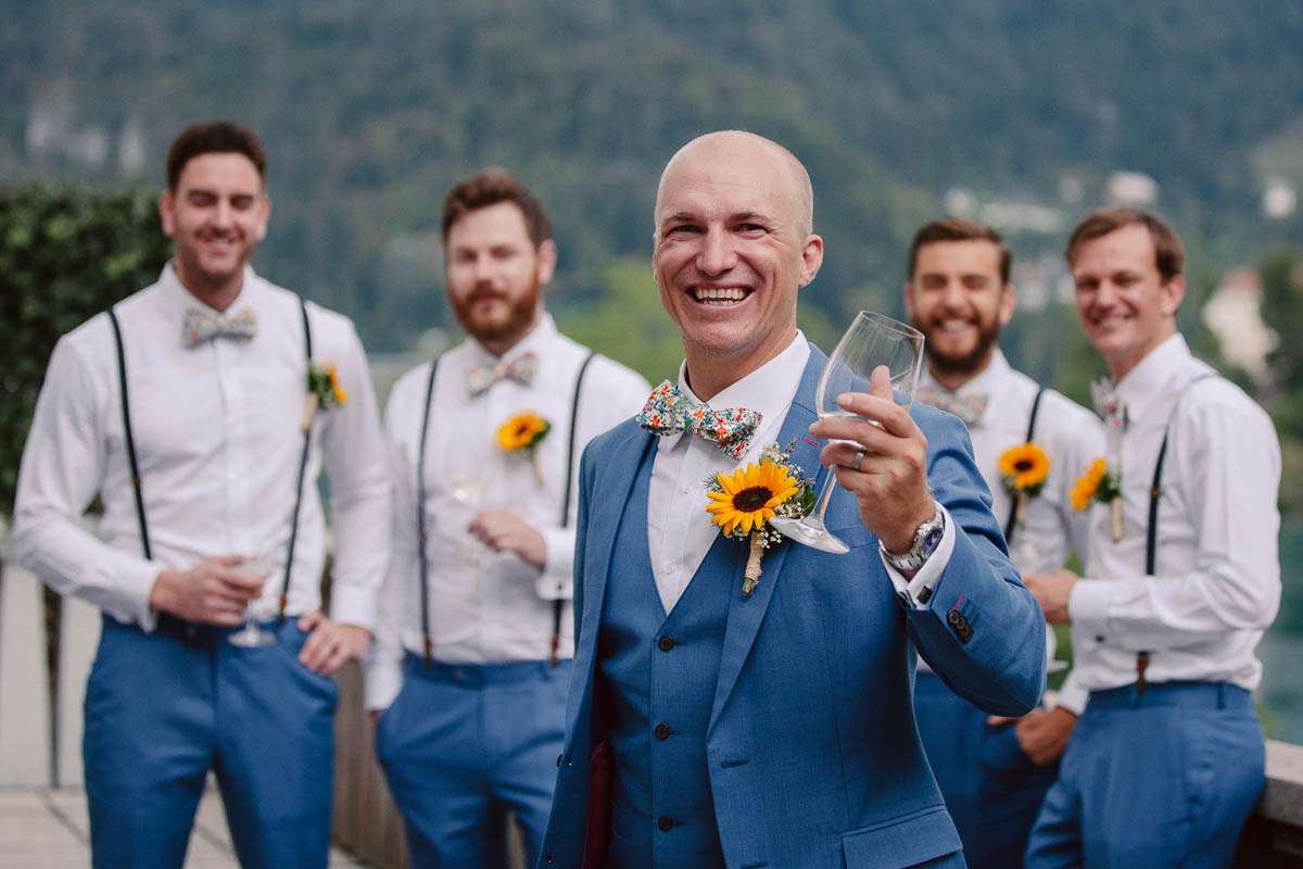 A groom in a blue suit holds a glass and smiles with his groomsmen standing behind him, all wearing matching outfits with sunflower boutonnieres, outside with greenery in the background.