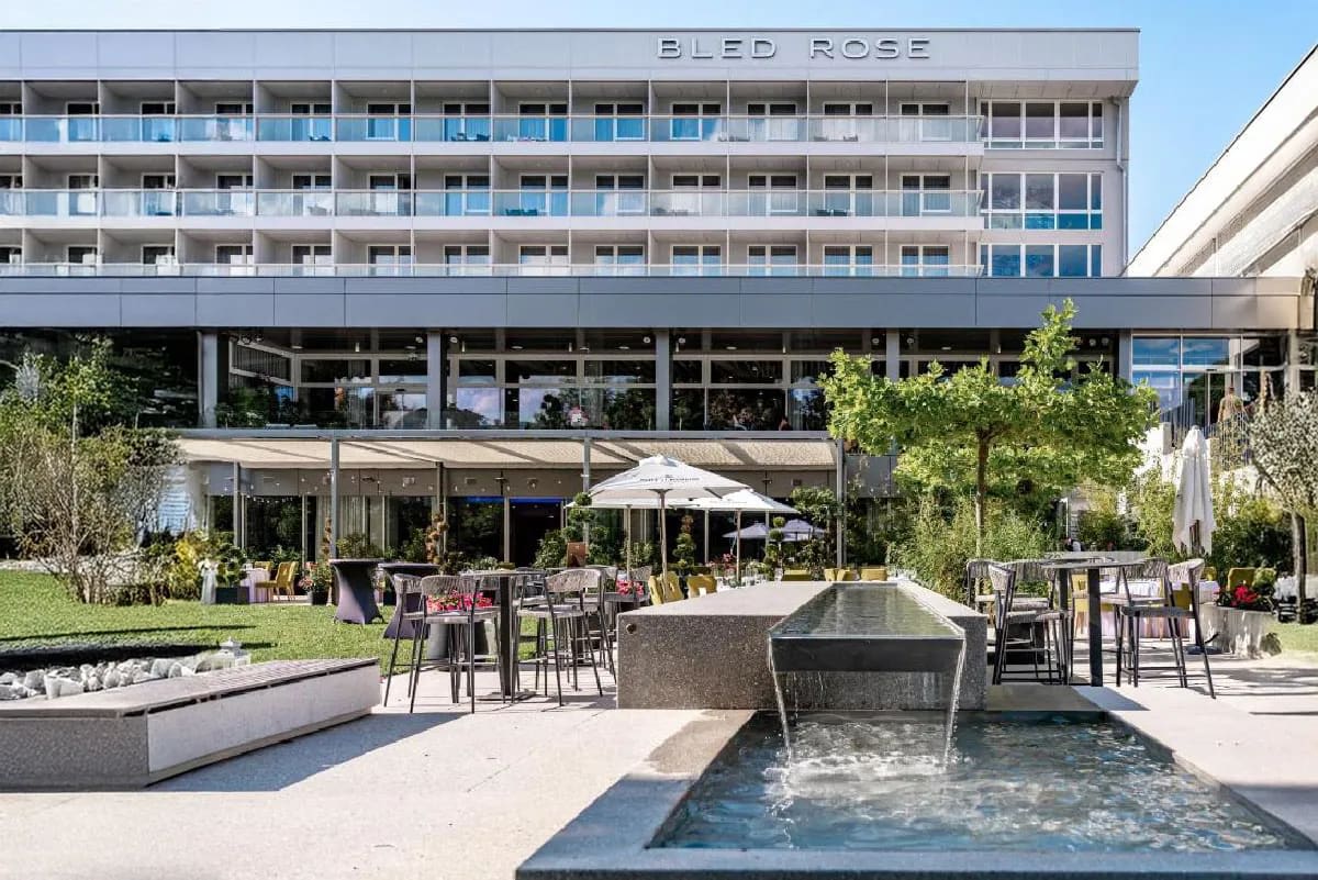 Outdoor seating area in a garden with a modern hotel in the background, featuring a small water fountain.