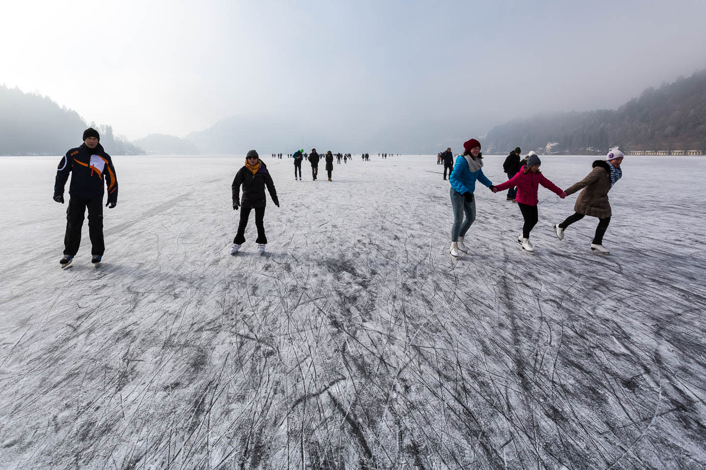 People enjoying ice skating on a frozen lake surrounded by snow-covered mountains and forested hills.