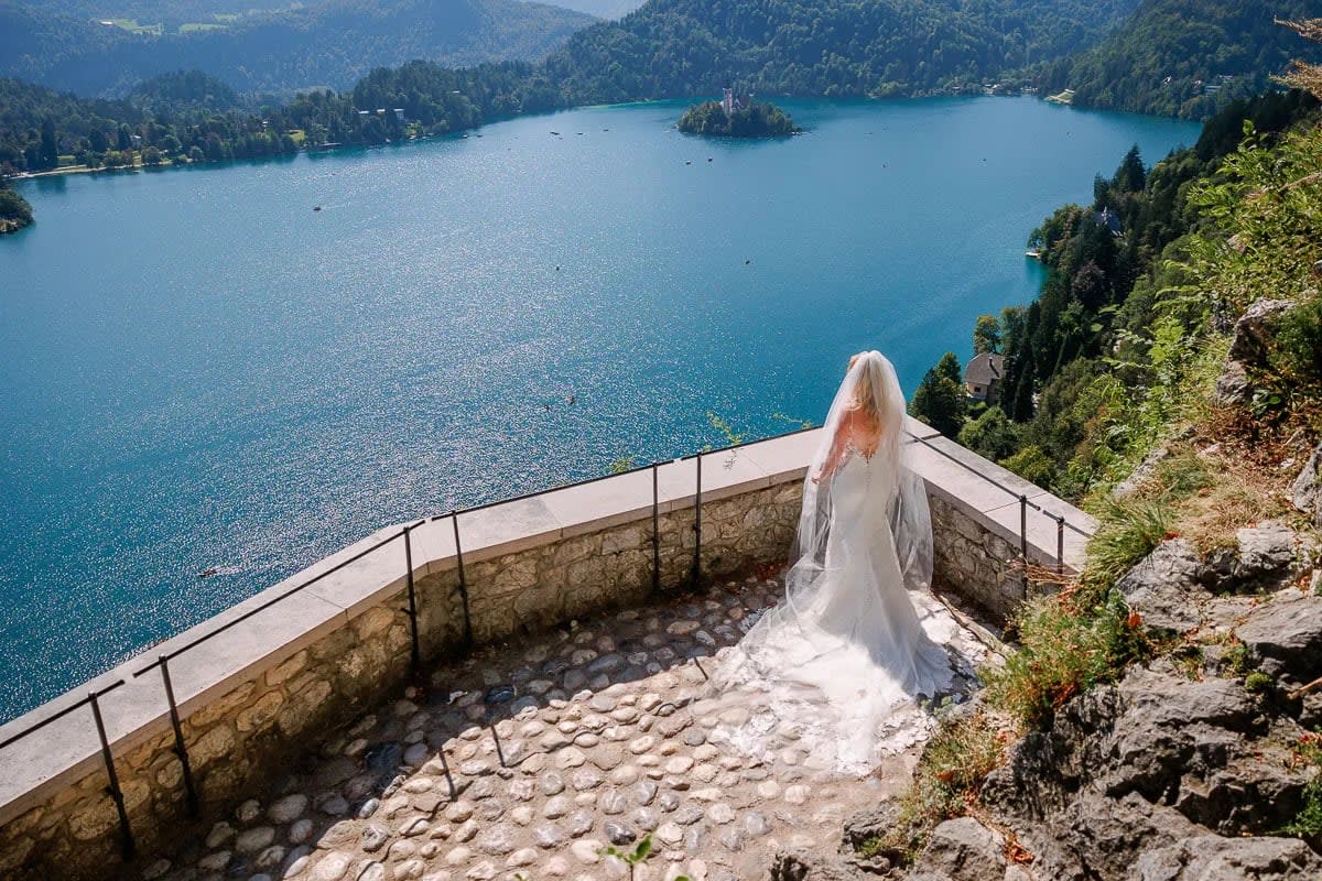 A bride in a wedding dress and veil stands on a stone terrace overlooking the stunning Lake Bled and its island on a sunny day.