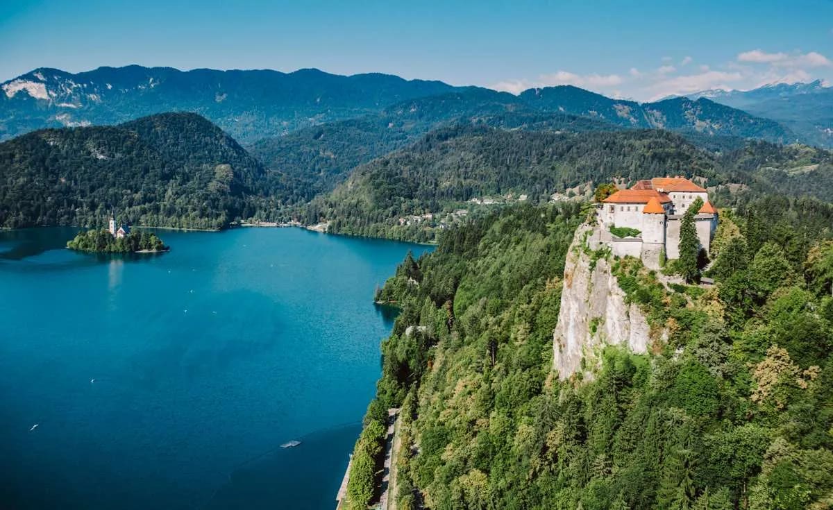 A scenic view of Lake Bled with a castle perched on a cliff and an island with a church surrounded by lush forests and mountains.