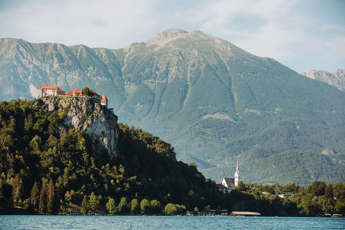 A scenic view of Lake Bled with a castle perched on a cliff and mountains in the background.