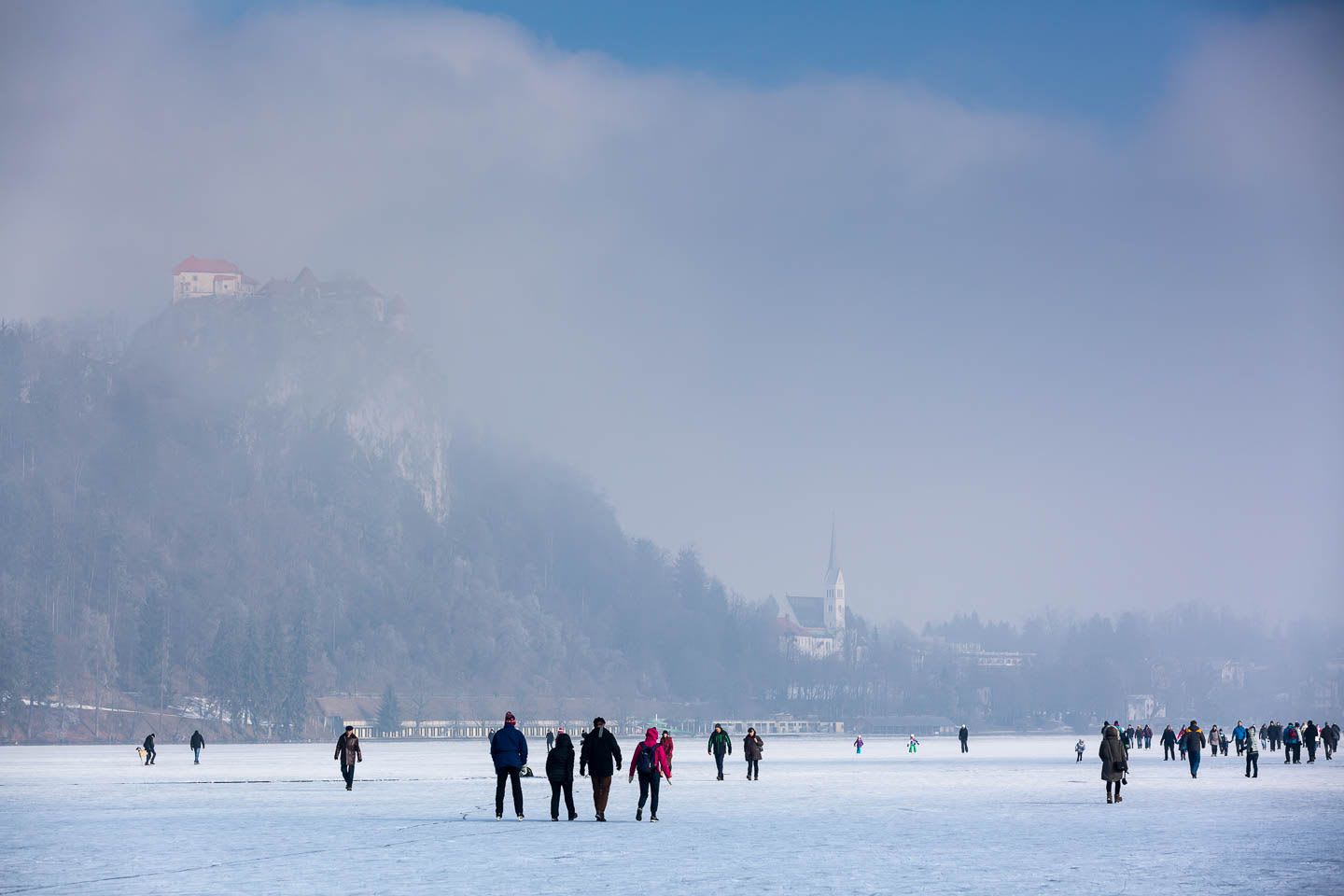 People walking on a frozen Lake Bled with a view of the castle perched on a hill and a church in the background on a misty day.