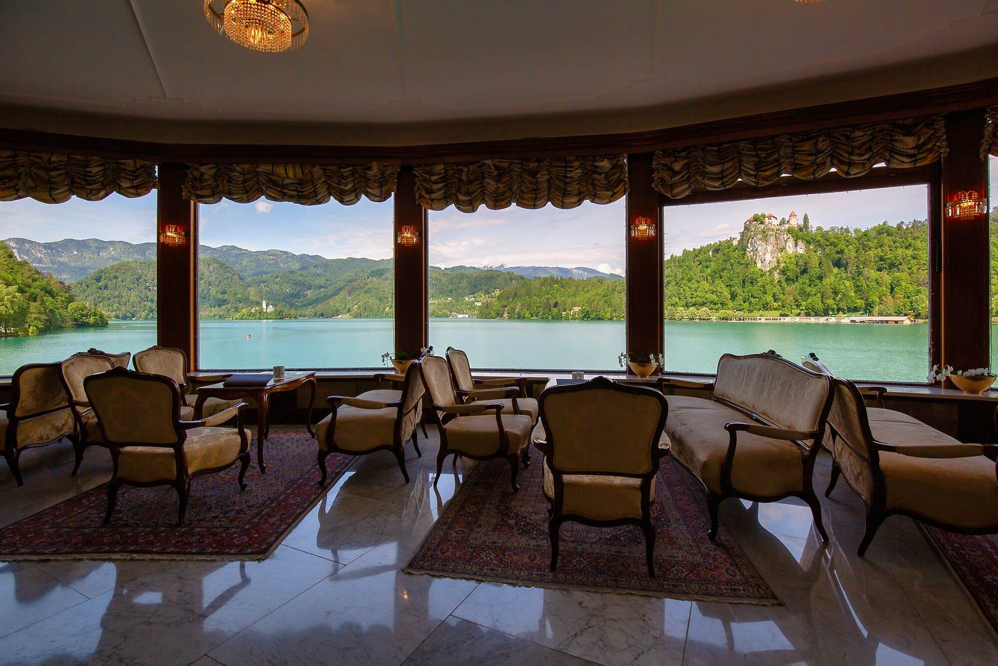 An elegant lounge with vintage chairs and large windows overlooking Lake Bled, featuring views of a castle and green mountains in the background.