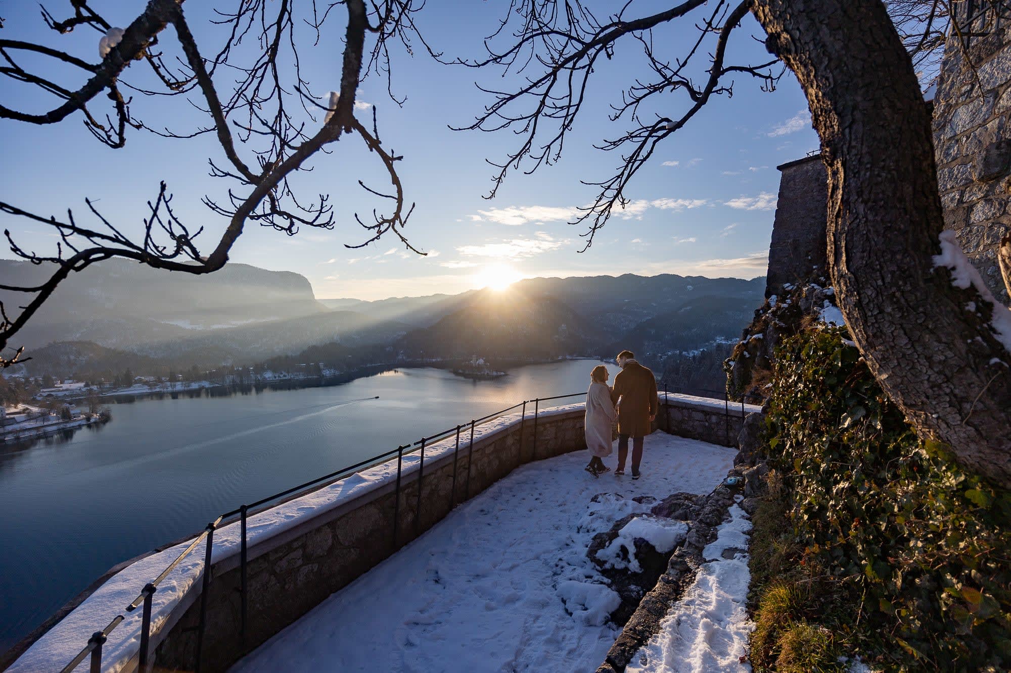 Couple walking on a snowy terrace at Bled Castle during winter sunrise, overlooking Lake Bled and the surrounding mountains.