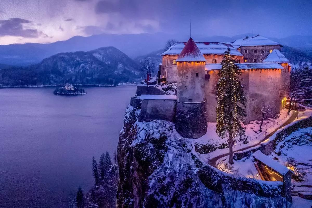 A winter view of Lake Bled with the illuminated Bled Castle on a snowy cliff and the island in the background.