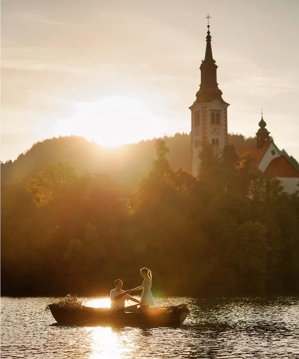 A couple stands on a boat during sunset with a church and hills in the background at Lake Bled.
