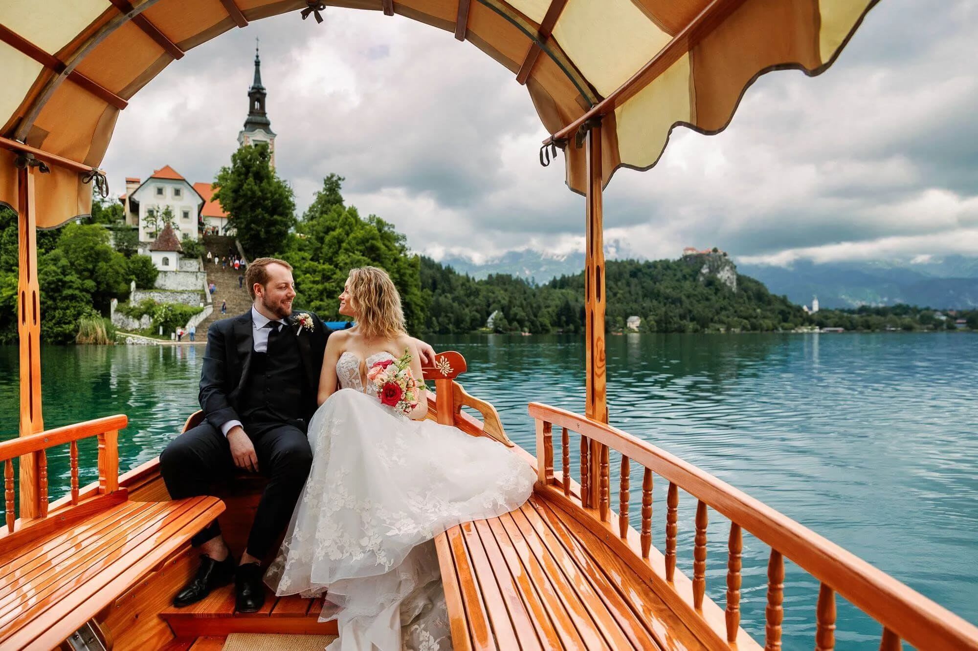 A bride and groom sitting together on a traditional wooden boat with the scenic view of Lake Bled and the island church in the background.