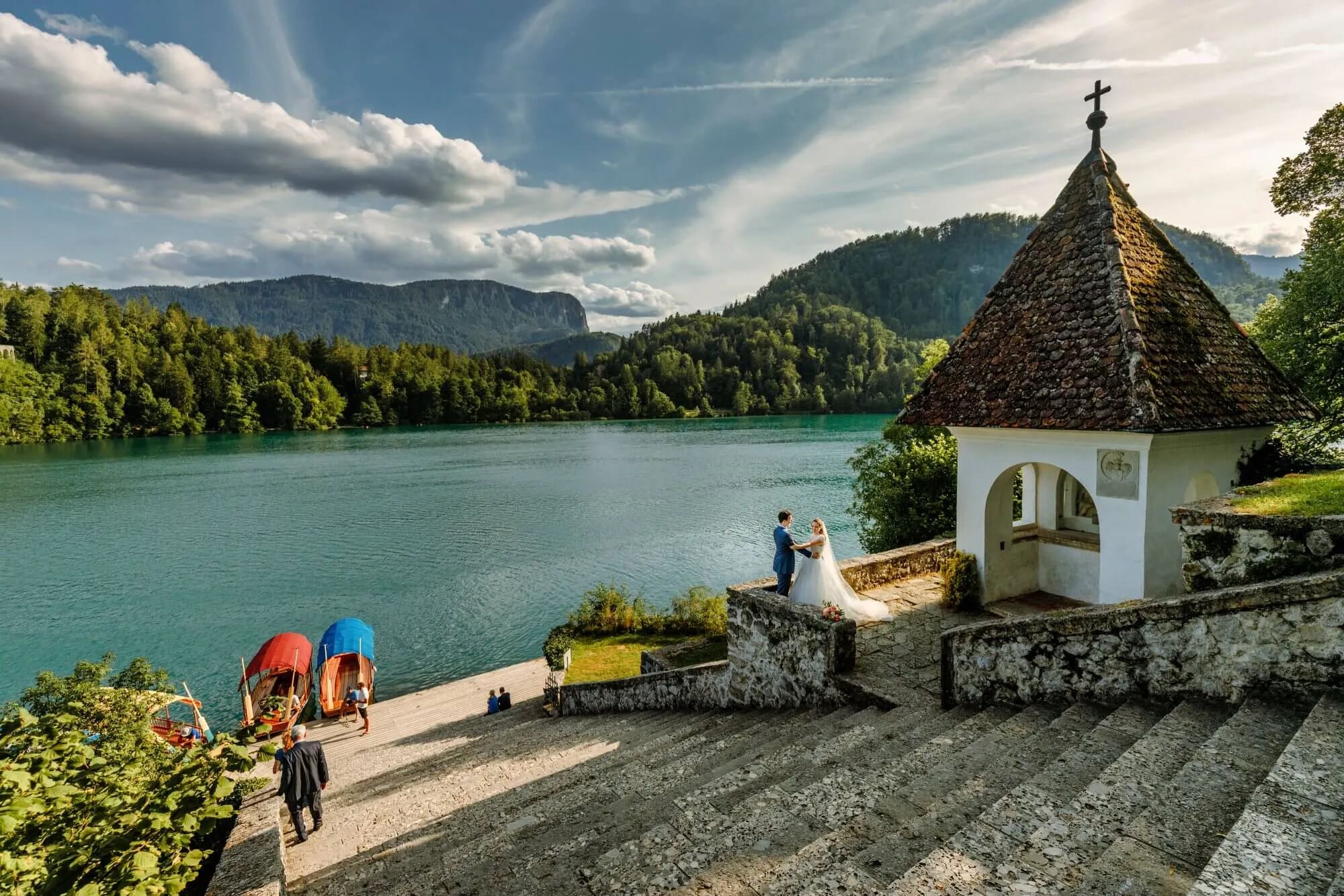 A couple holding hands by a small chapel, overlooking the serene Lake Bled with mountains and boats in the background.