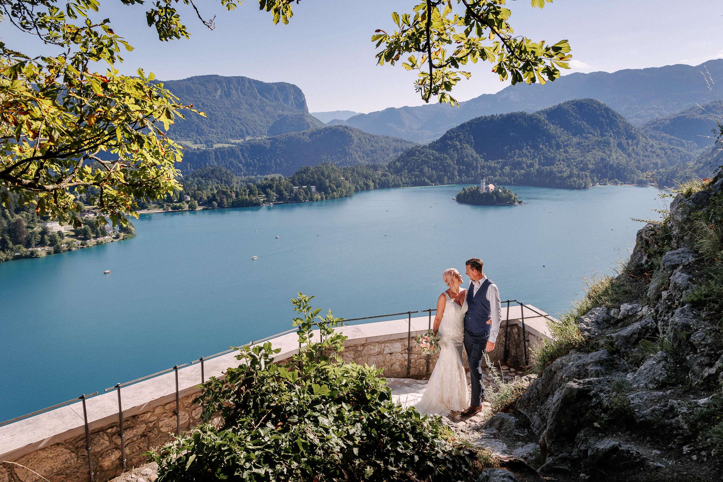 Bride and groom standing on a terrace overlooking Lake Bled with Bled Island and the mountains in the background.