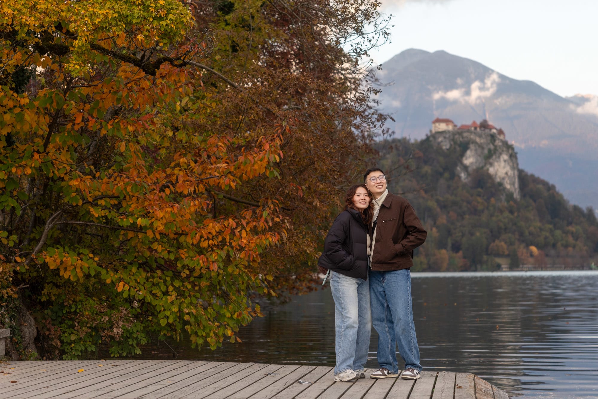 ouple standing on a dock with autumn colours and Bled Castle in the background at Lake Bled.