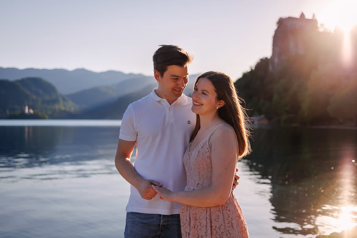 A couple smiling at each other by the water with a view of a castle on a hill at Lake Bled.