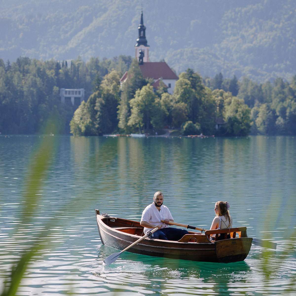 A couple enjoying a ride in a wooden rowboat on Lake Bled, with a church and lush greenery in the background.