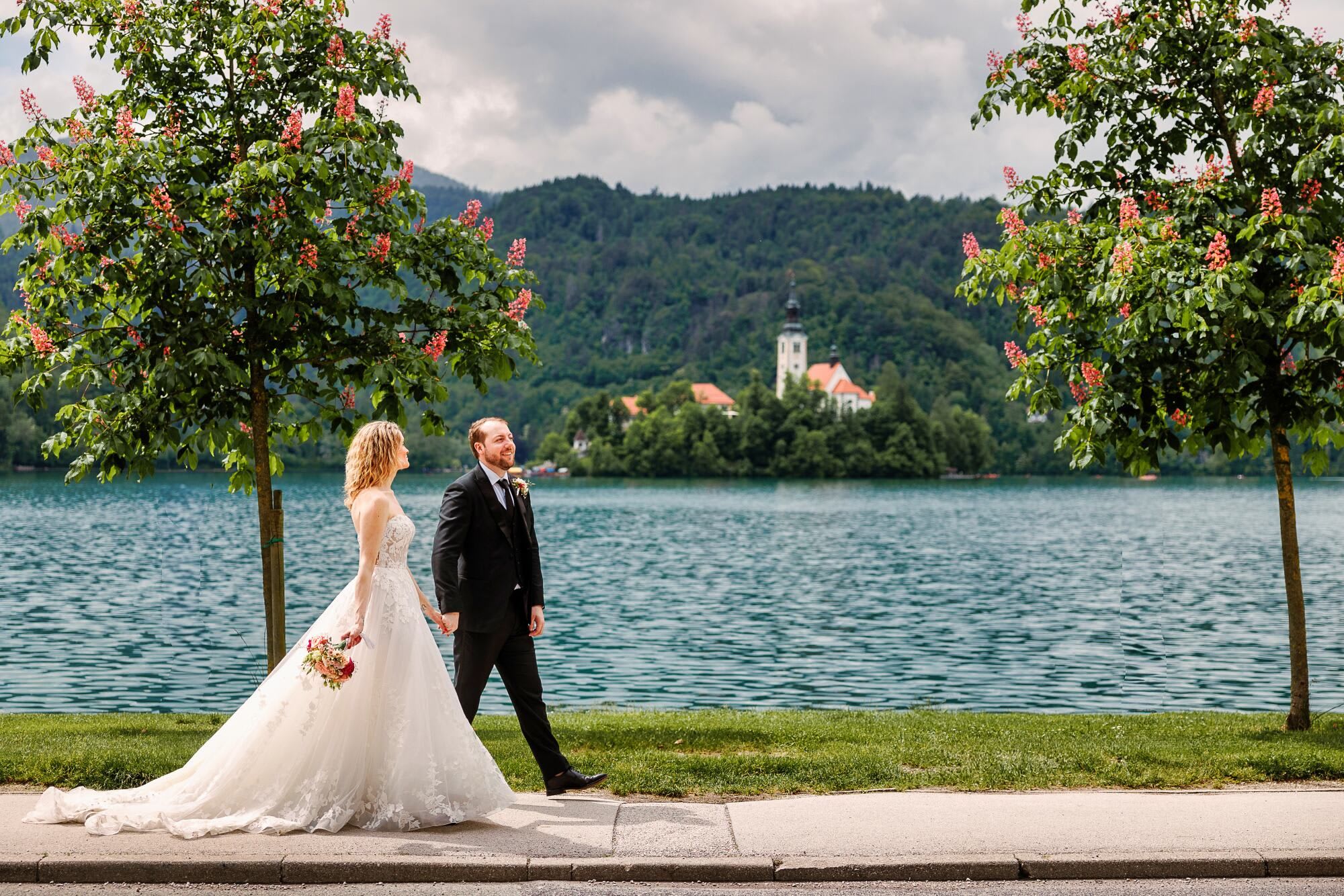 A bride and groom holding hands and walking along the lakeside path with the church in the background at Lake Bled.
