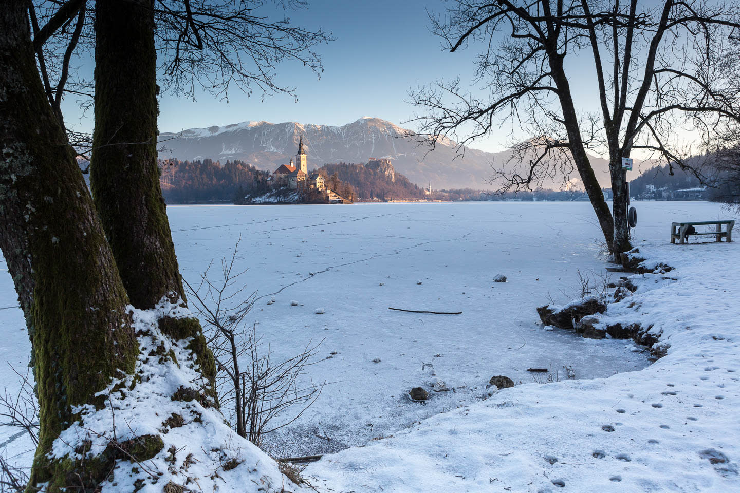 A winter scene at Lake Bled with a view of the island church, surrounded by frozen waters and snow-covered landscape.