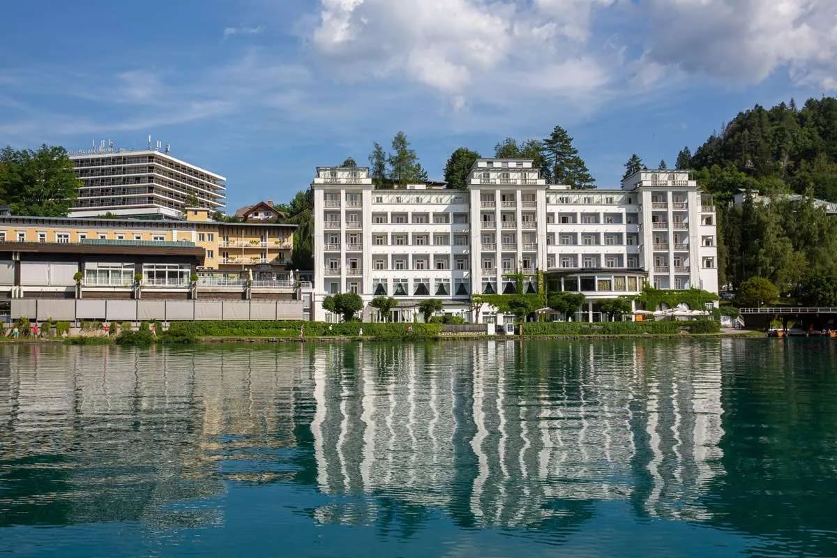 A view of a grand hotel on Lake Bled's waterfront with clear reflections on the water and a bright blue sky.