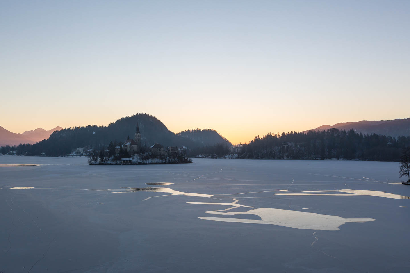 A serene view of Lake Bled with its frozen surface, the island church, and the surrounding mountains at sunset.