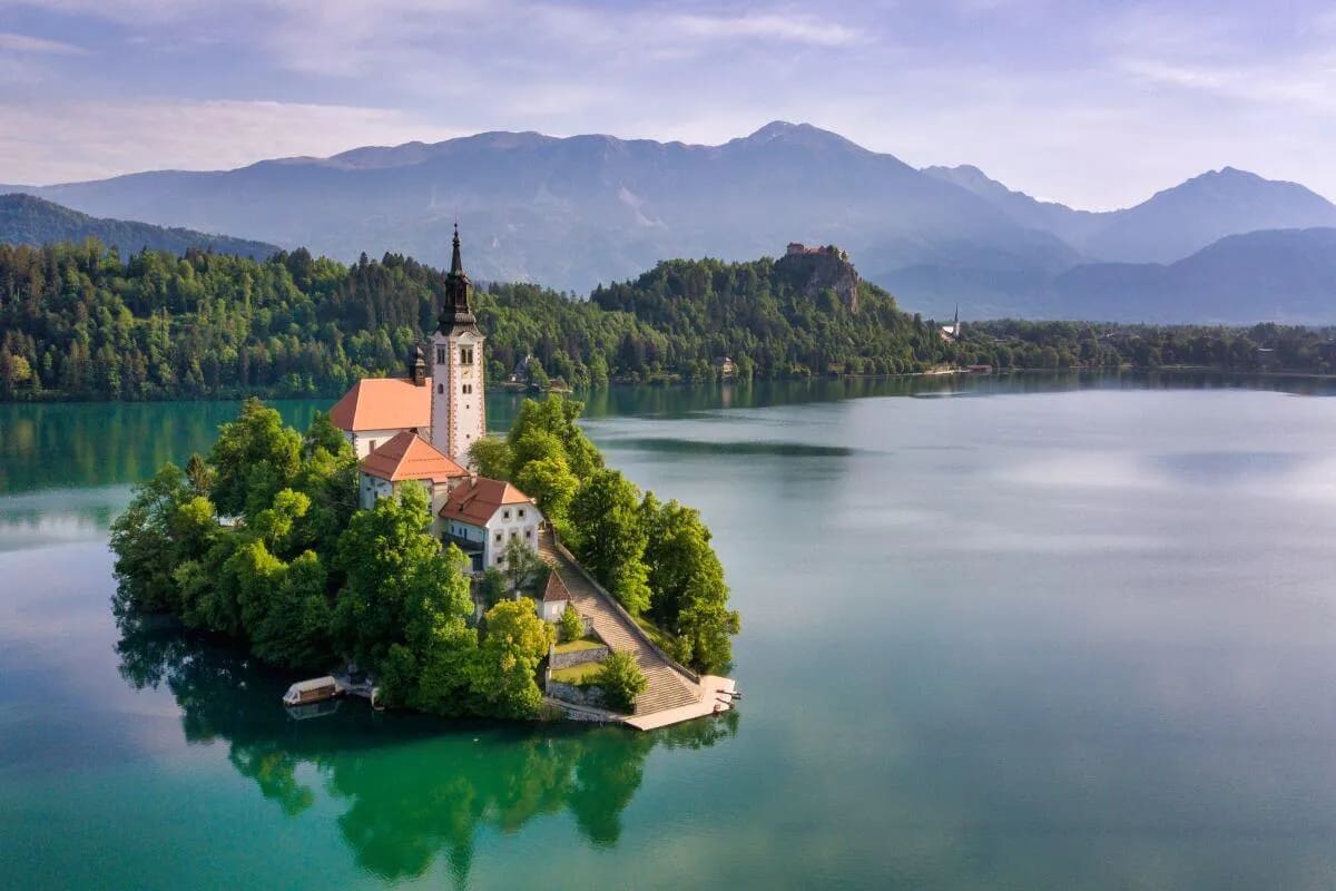 A serene view of the church on Bled Island surrounded by clear blue water and lush greenery, with mountains in the background.
