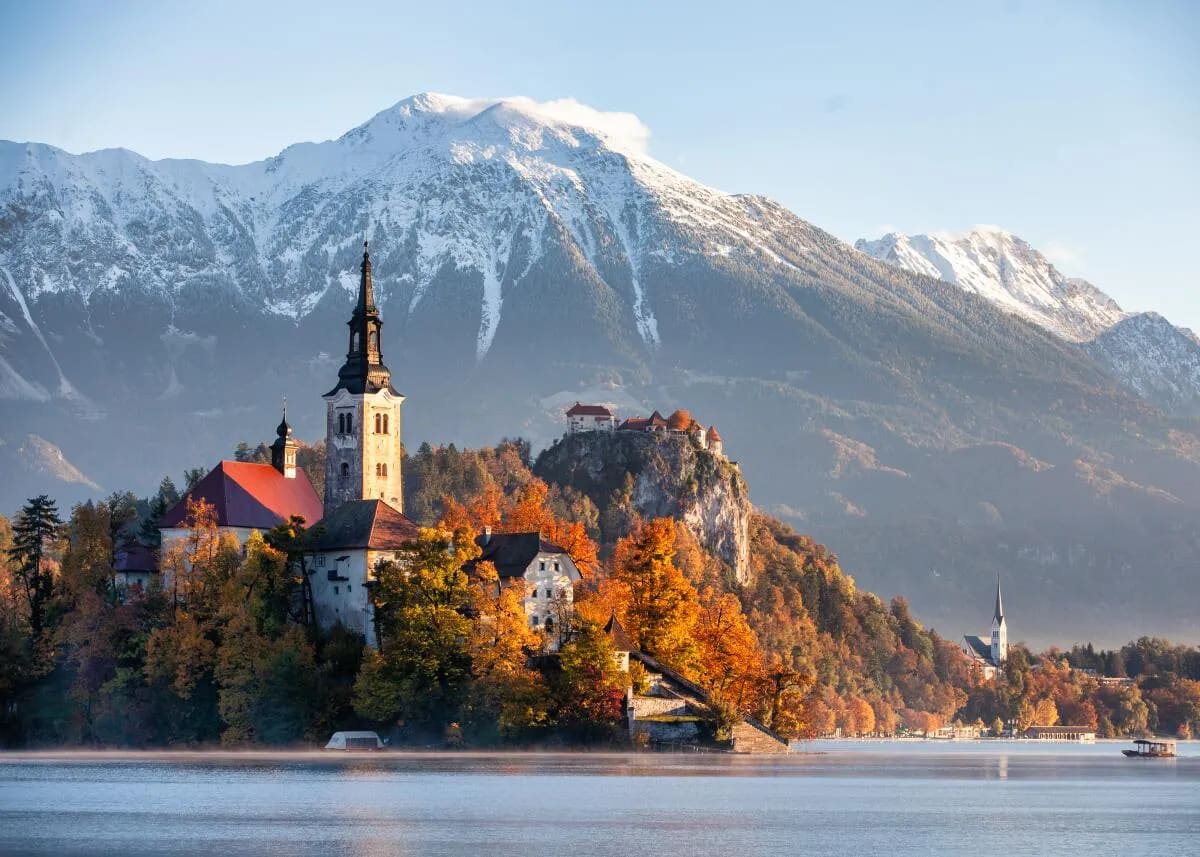 A scenic view of Lake Bled with its iconic island church, surrounded by autumn foliage and snow-capped mountains in the background.