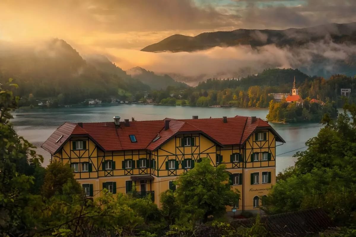 A picturesque view of Lake Bled with an island church surrounded by misty mountains and a charming yellow building in the foreground.
