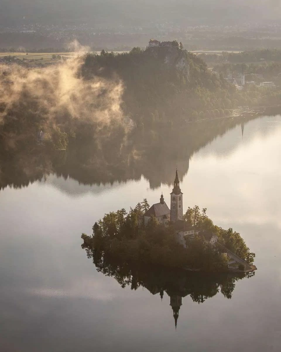 An aerial view of Lake Bled with its iconic island and church reflecting in the calm water, surrounded by mist and distant hills.