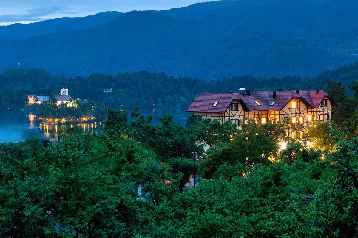 A scenic view of Lake Bled at dusk, showing the illuminated island with its church and the mountains in the background.