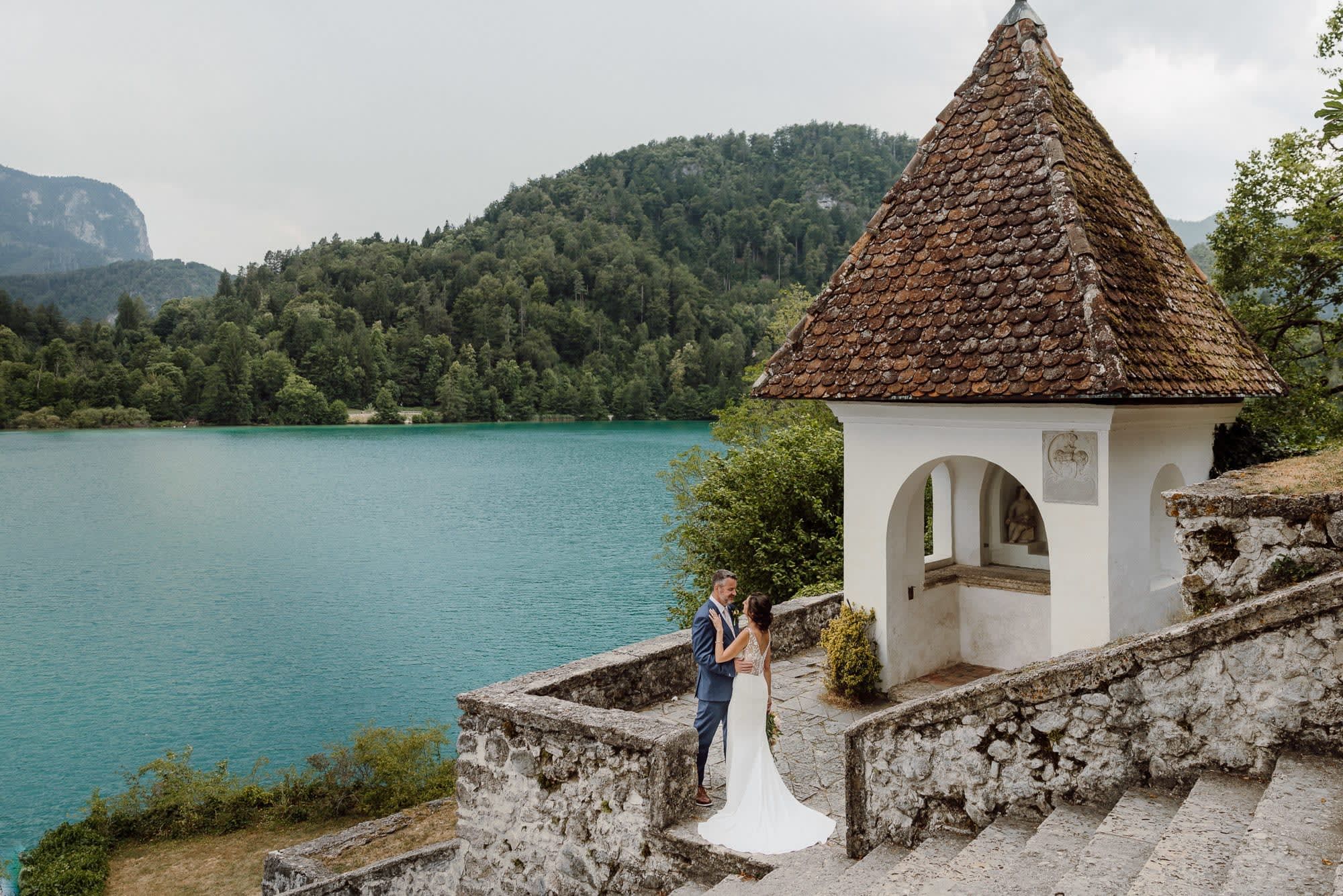 Bride and groom standing beside a small chapel on Lake Bled island with turquoise water and forested hills in the background.