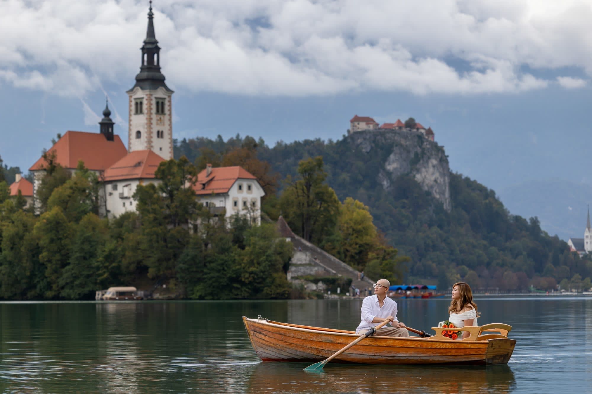 Post-wedding couple portrait on a wooden rowboat with Bled Island and Bled Castle in the background.