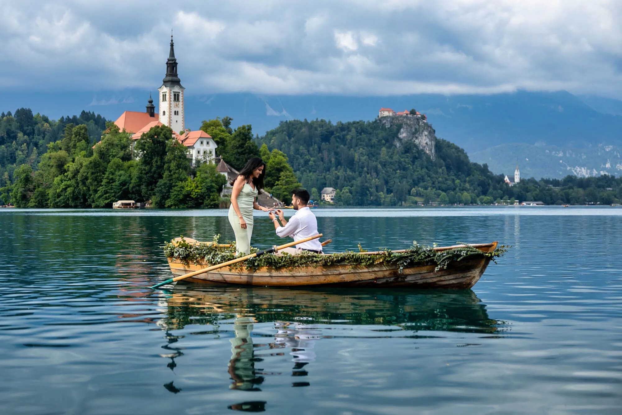 A couple in a boat on Lake Bled, with the man proposing to the woman. The church on the island, Bled castle and mountains are visible in the background.