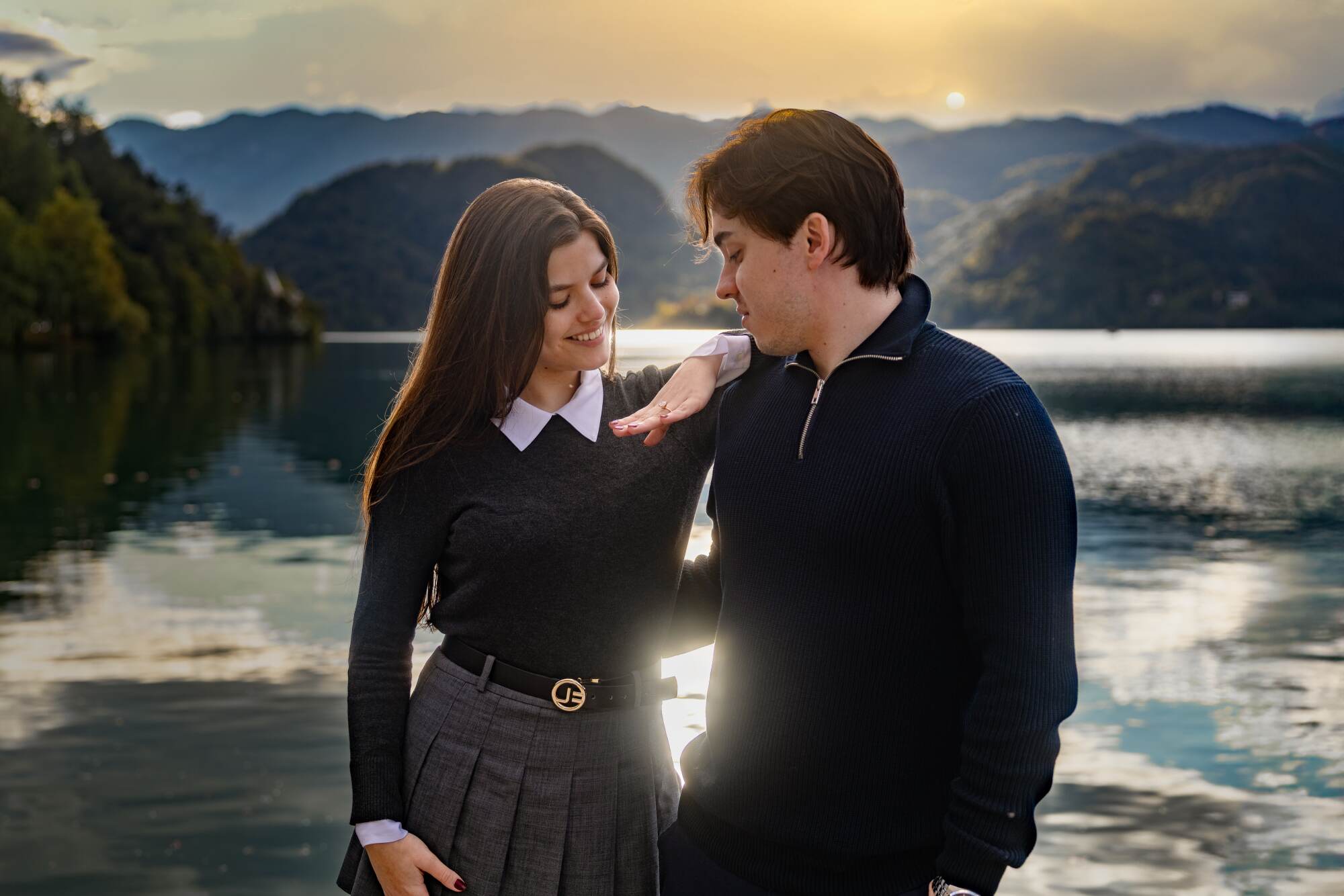 A newly engaged couple at Lake Bled, with the woman smiling while showing her engagement ring to her partner against a scenic mountain and lake backdrop.