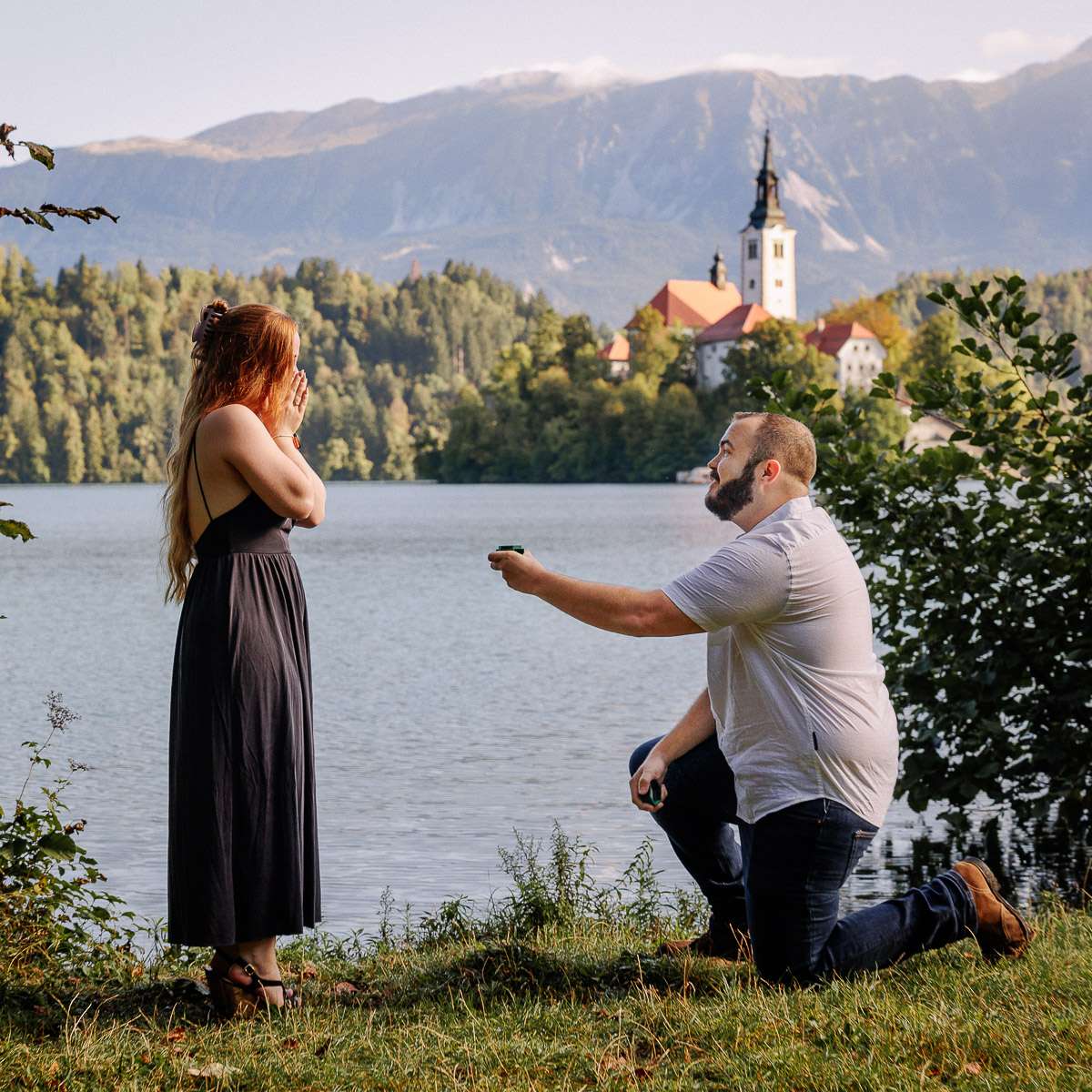 A man proposing to a woman by the lake with a view of the island church and mountains in the background.