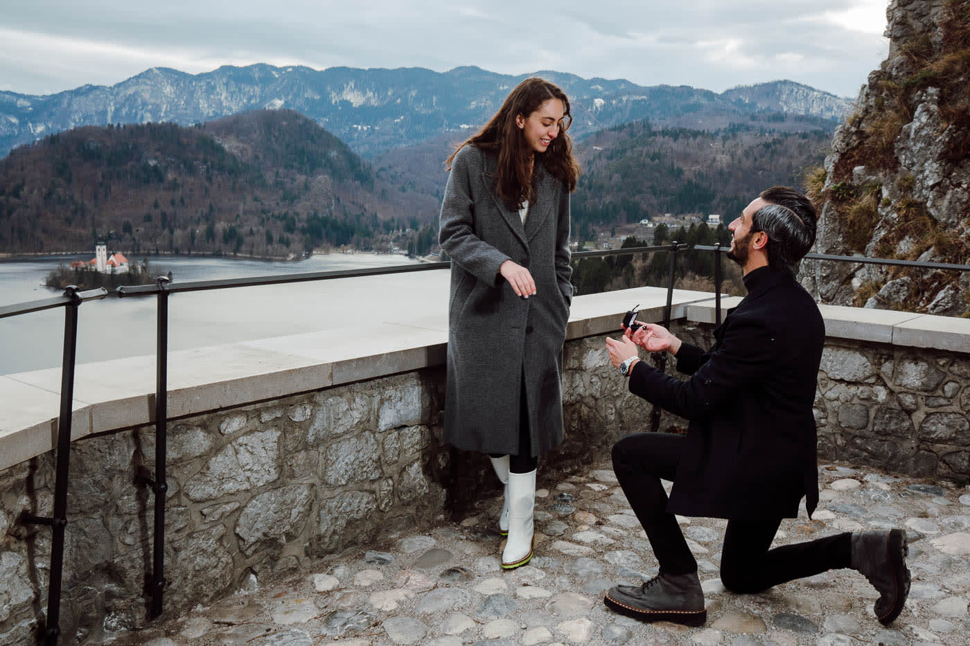 A man kneeling to propose to a woman on a scenic terrace, with Lake Bled and the mountains visible in the background.