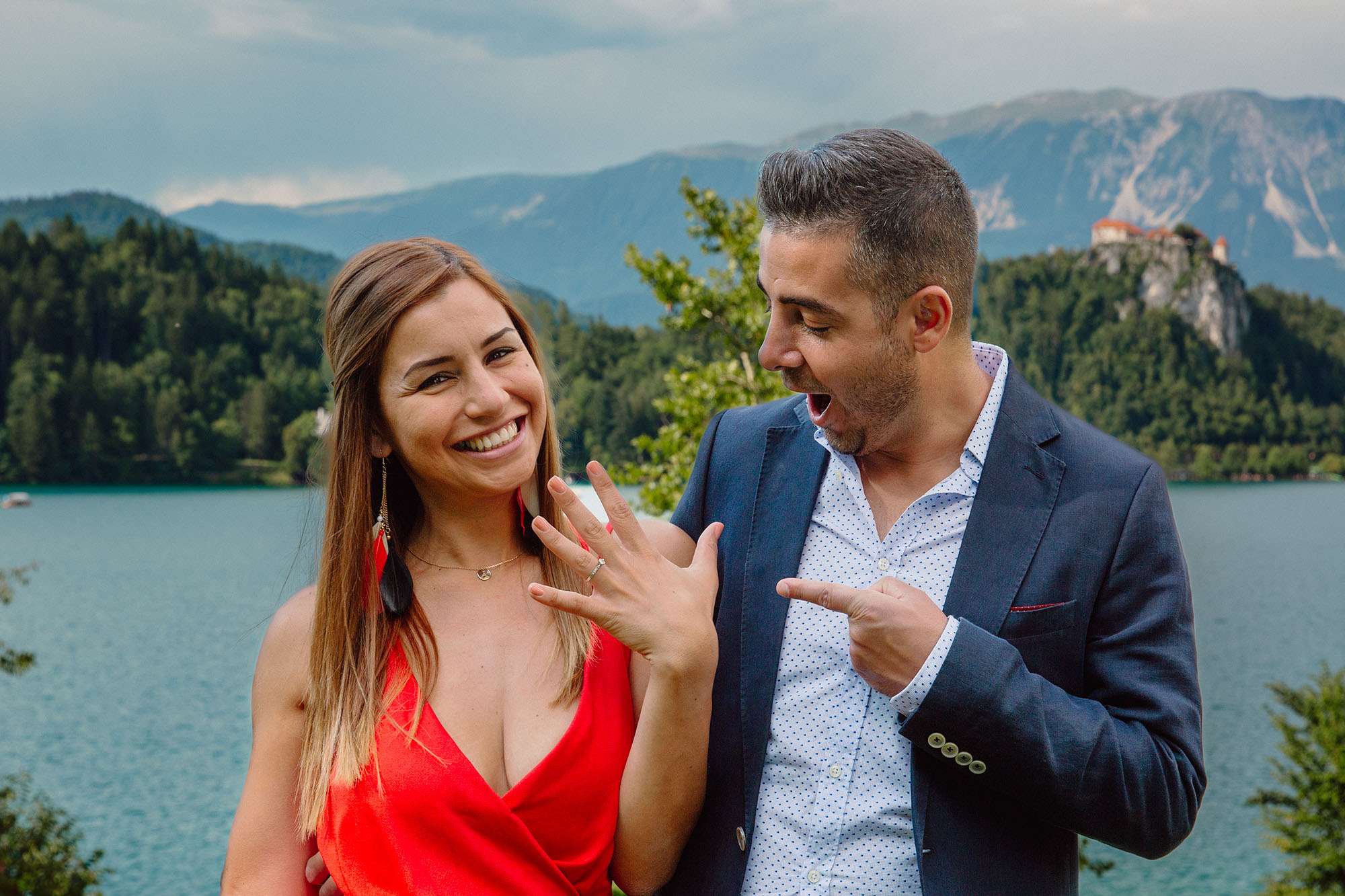 A happy couple celebrating a marriage proposal by a lake, with the man excitedly pointing at the woman's engagement ring and a scenic view of mountains and a castle in the background.