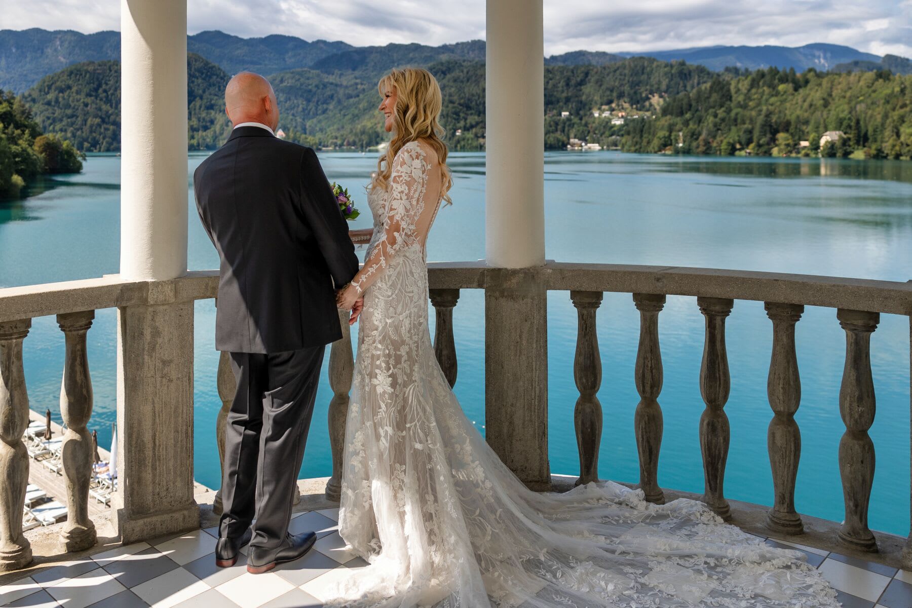 A bride and groom standing on a balcony overlooking the serene waters of Lake Bled with mountains in the background.
