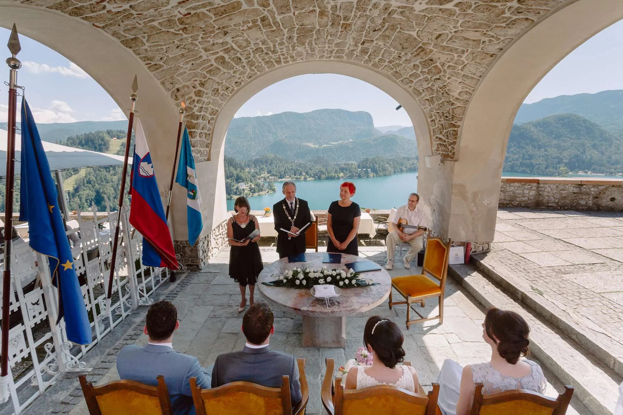 A wedding ceremony taking place under an arched structure on Bled castle with views of Lake Bled and the surrounding mountains in the background.