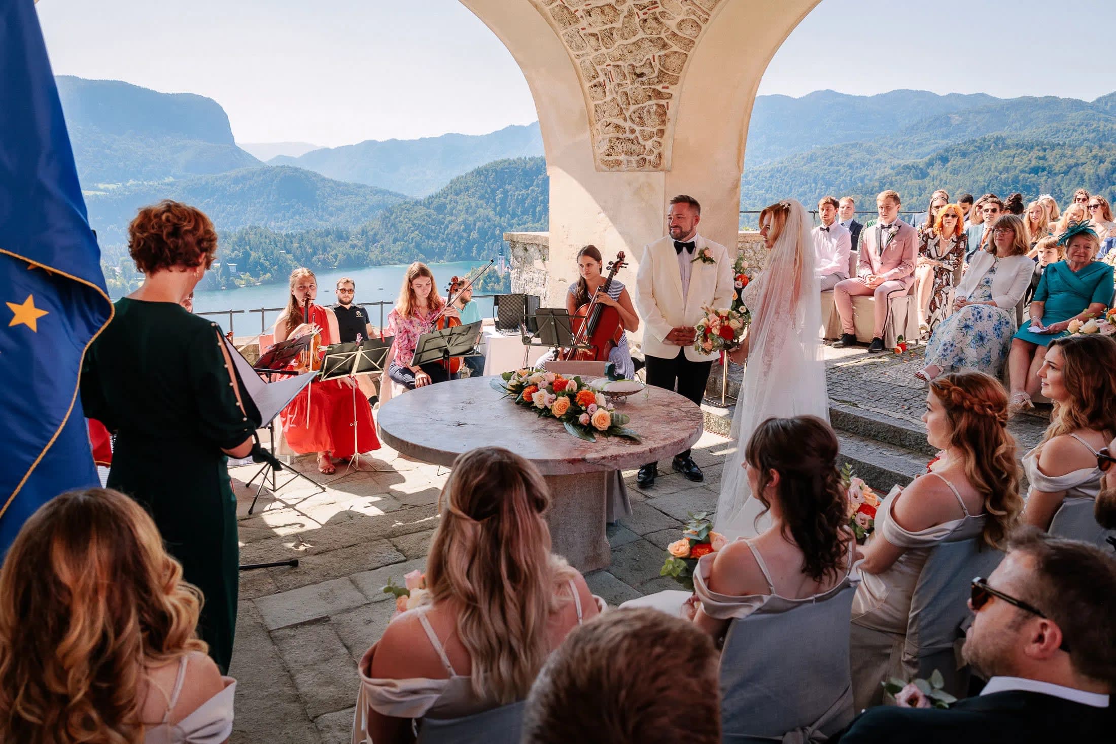 A wedding ceremony with the bride and groom standing under an arch, accompanied by musicians and guests overlooking a scenic view of Lake Bled and surrounding mountains.