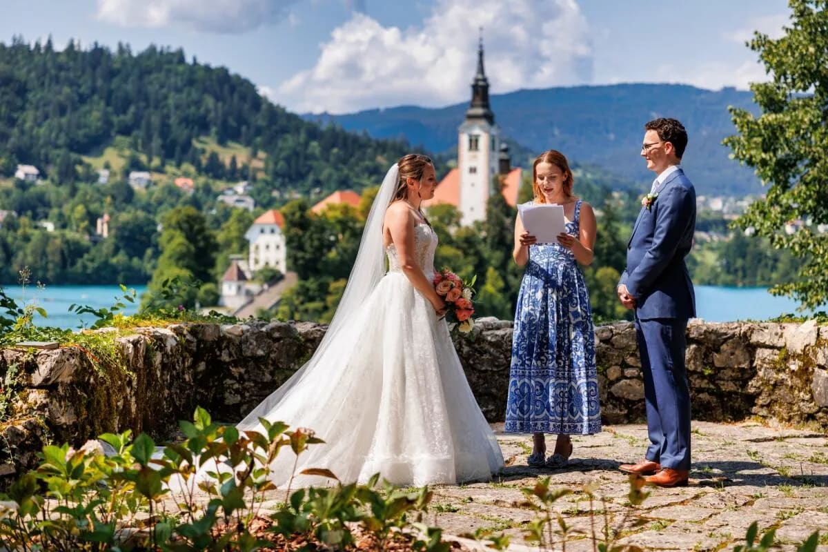 A bride and groom exchanging vows outdoors with a view of Lake Bled and a church in the background.