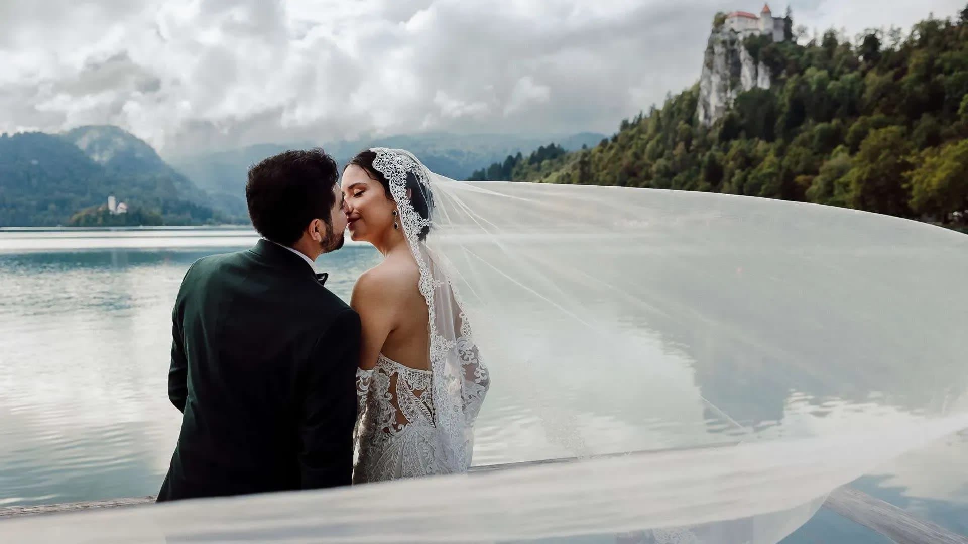 Bride and groom sharing a kiss by Lake Bled with a flowing lace veil, Bled Castle and the island church in the background.