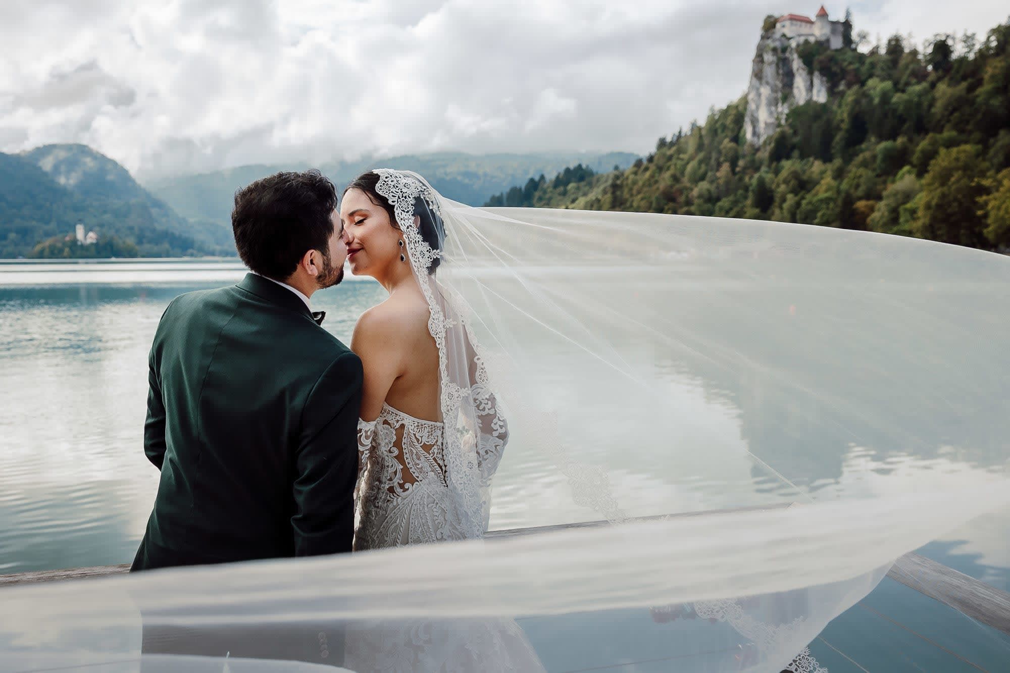 Bride and groom sharing an intimate moment by Lake Bled, with flowing veil and Bled Castle in the background.