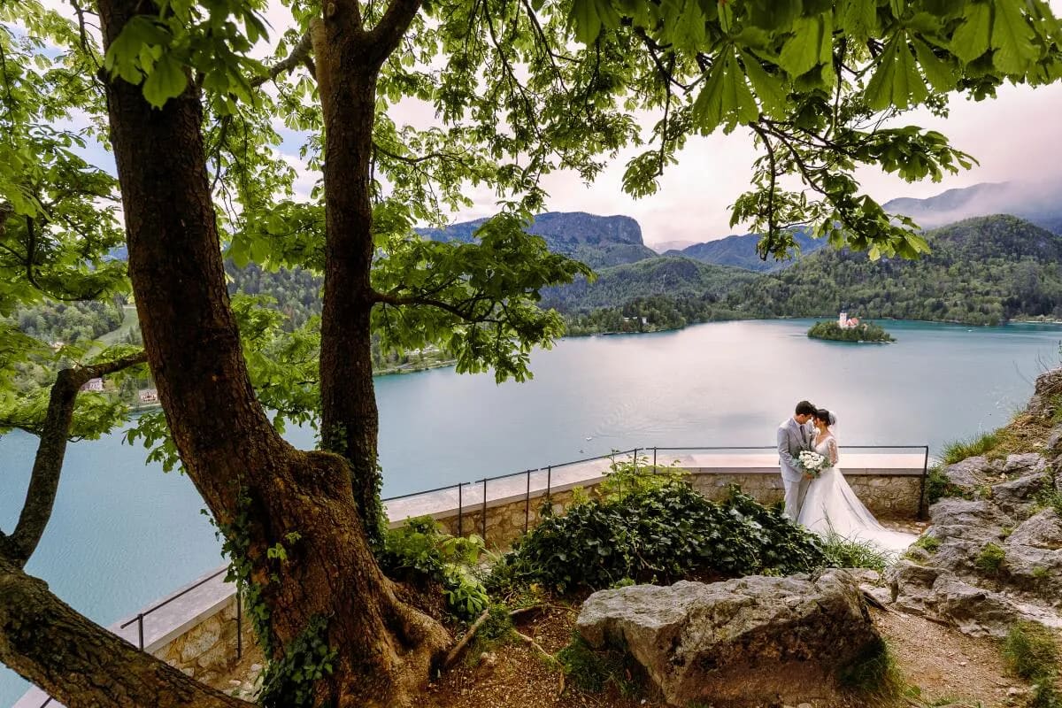 A couple embracing on a terrace with a scenic view of Lake Bled and its island in the background, surrounded by lush greenery.