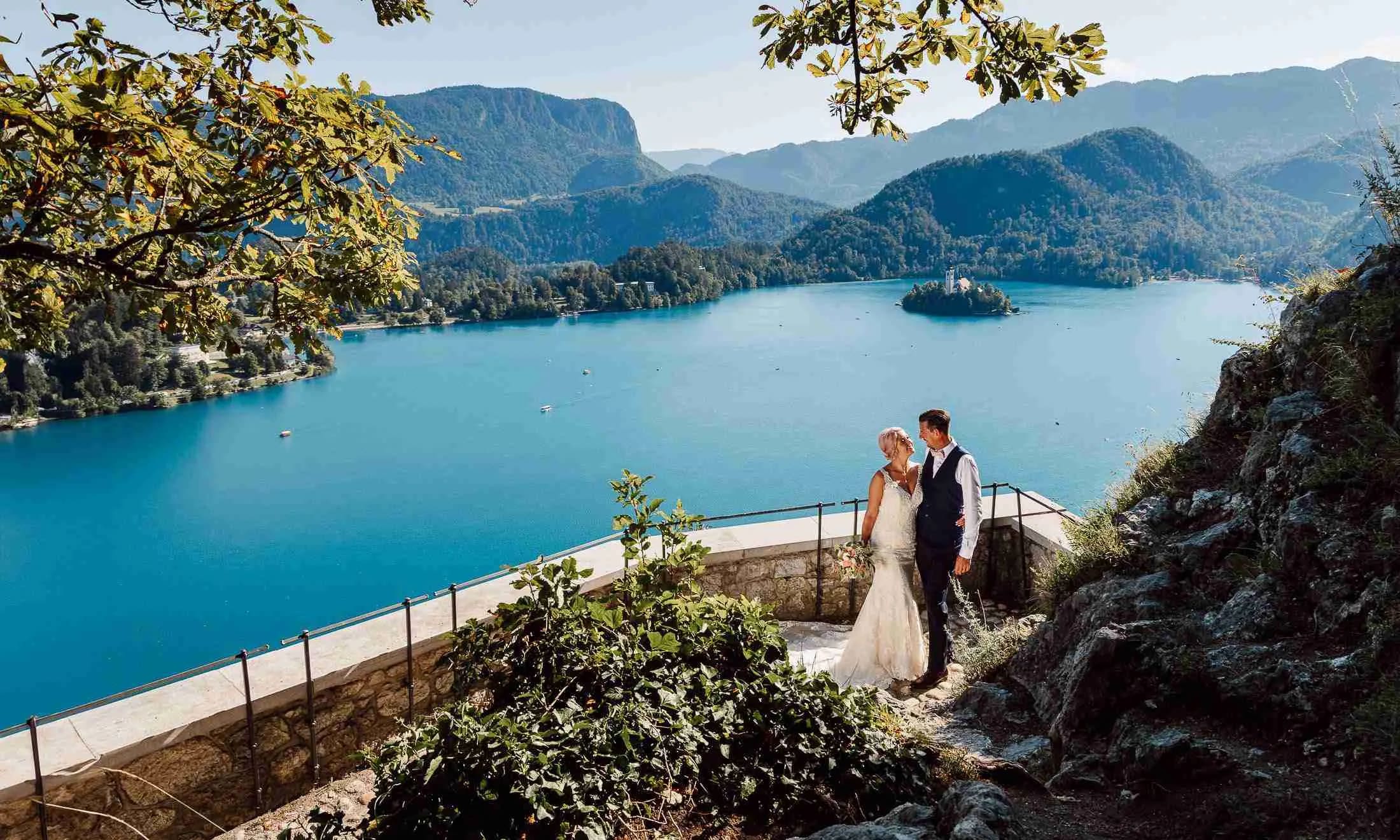 A bride and groom standing on a viewpoint overlooking Lake Bled, with the iconic island and mountains in the background.