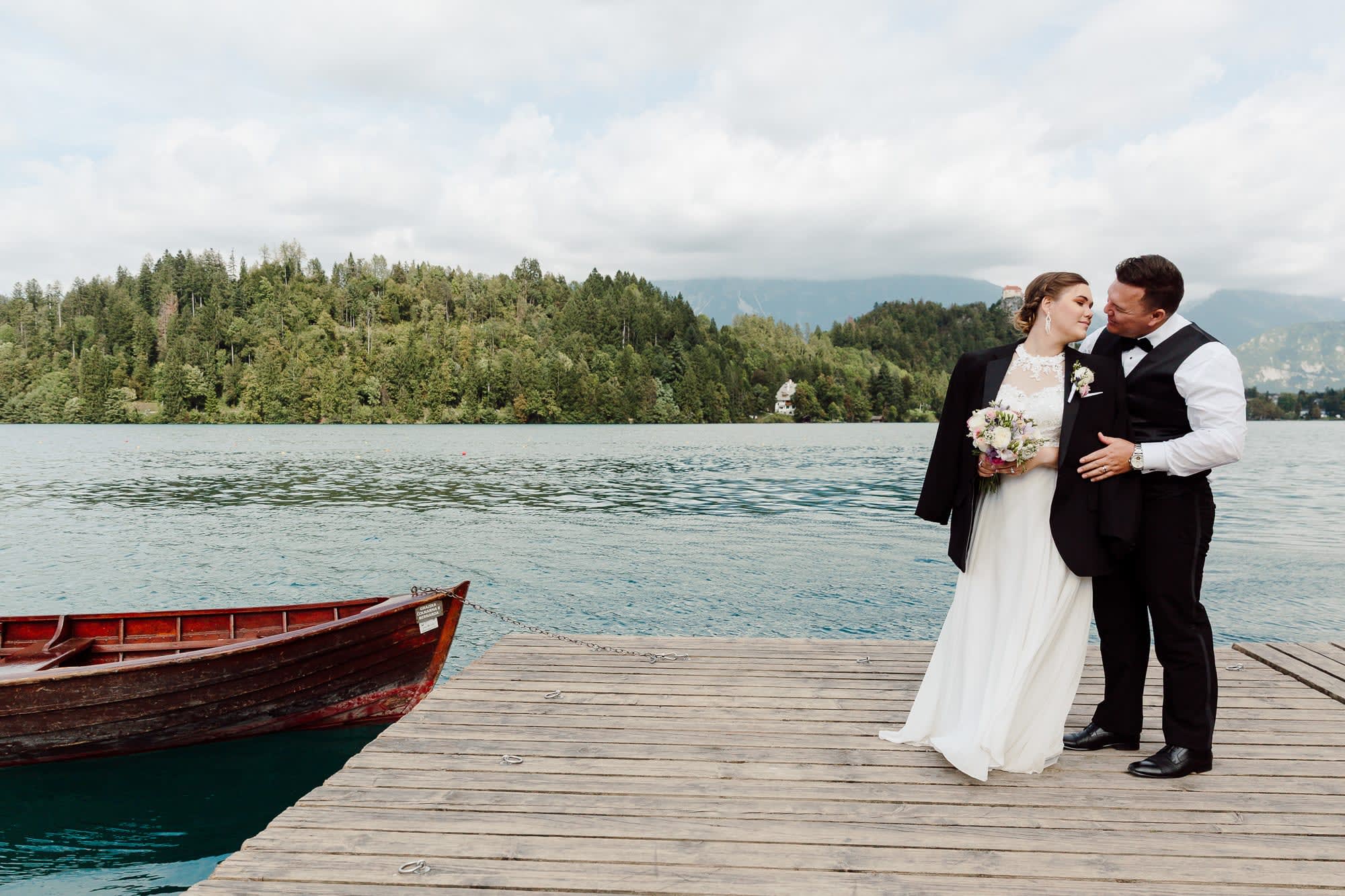 A bride and groom standing together on a wooden dock beside a boat, with Lake Bled and forested hills behind them.