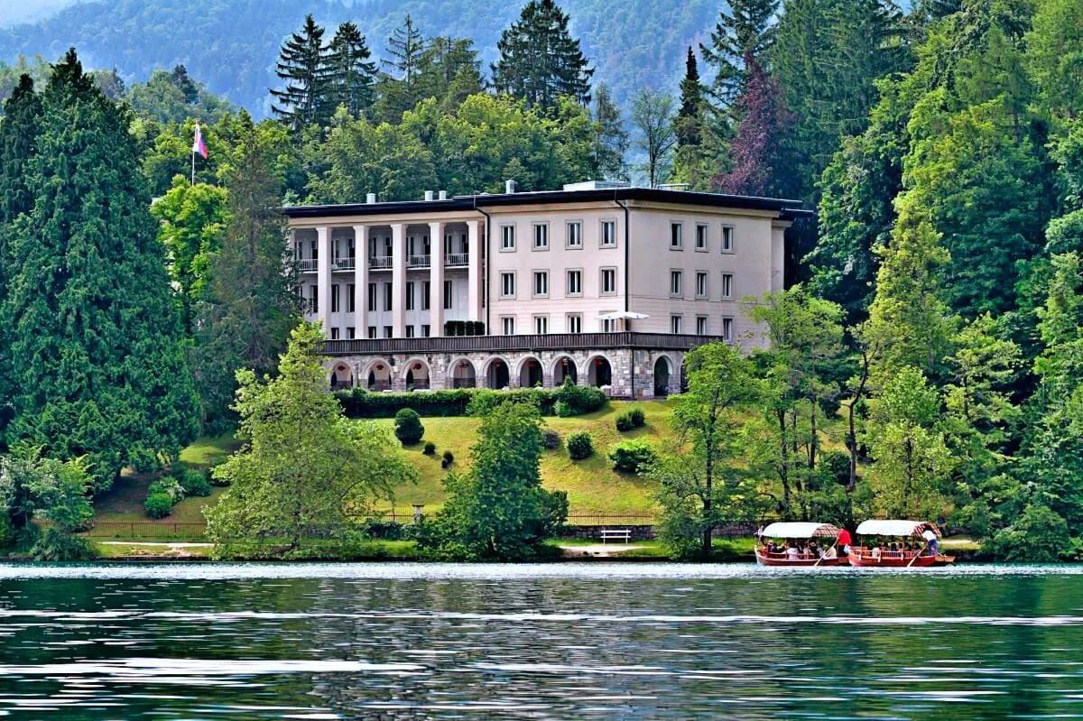 A large elegant building surrounded by lush greenery near the water's edge with boats on Lake Bled below.