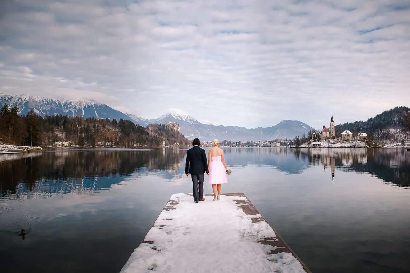 A couple holding hands and walking on a snowy dock with Lake Bled and its church island in the background.