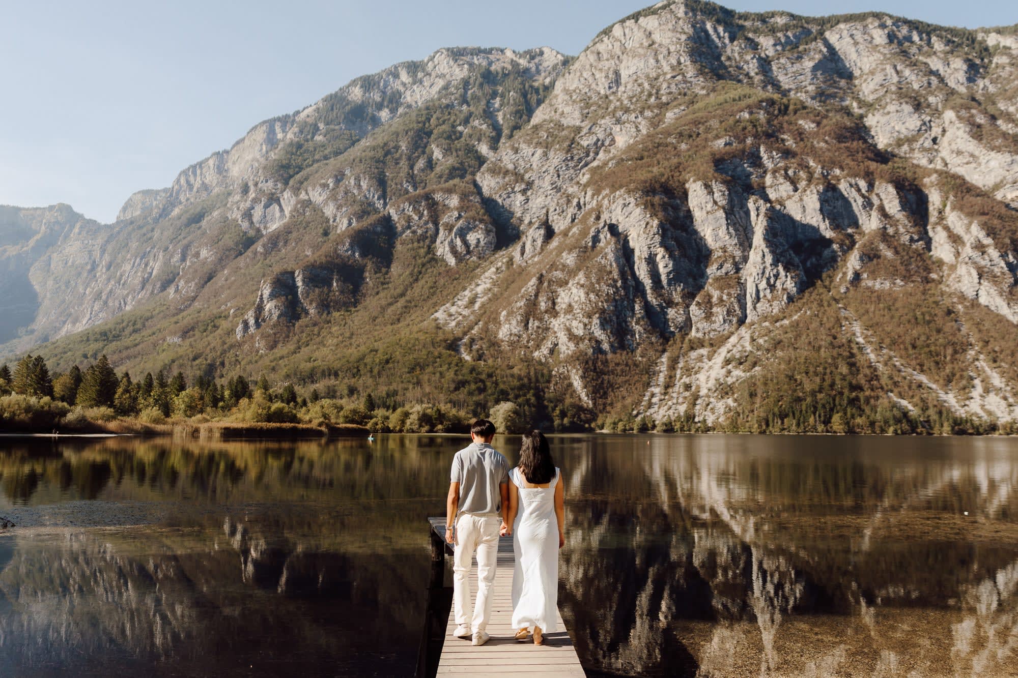 Couple walking hand in hand along a wooden jetty on Lake Bohinj, with the Julian Alps reflected in still water.