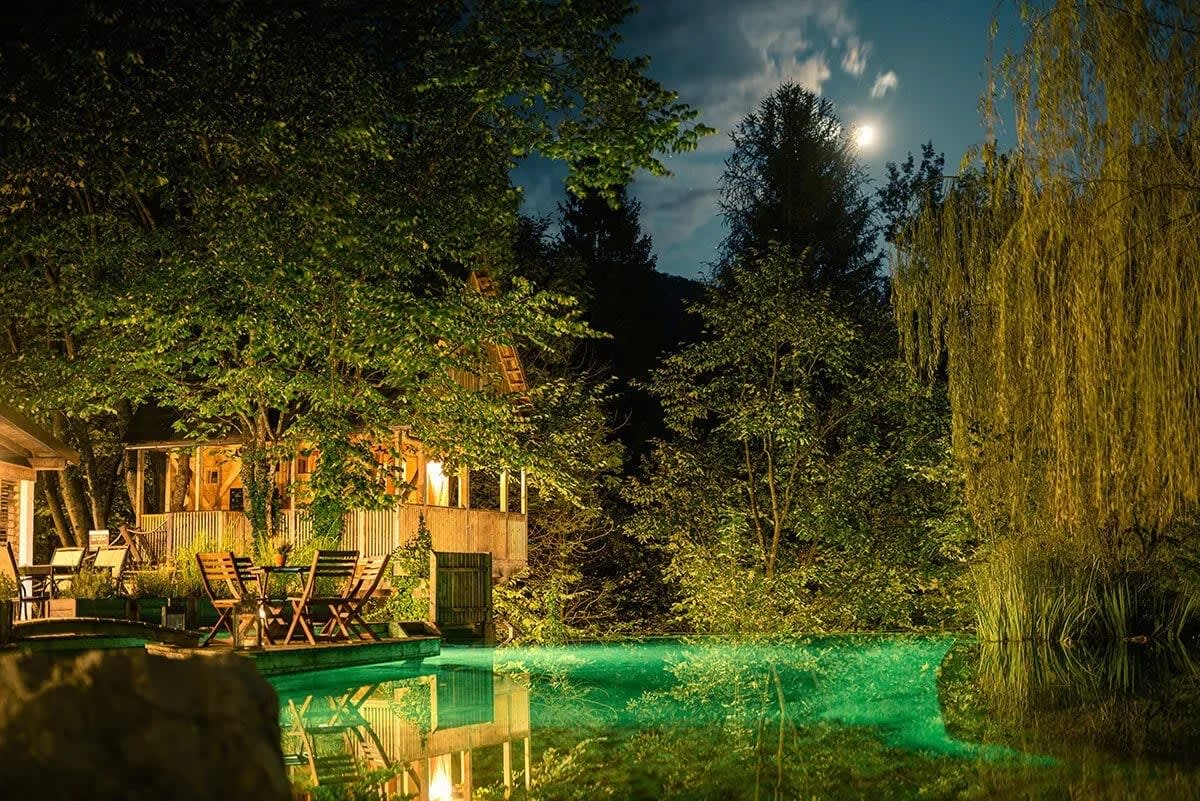 A cozy cabin surrounded by trees is beautifully illuminated at night, with a full moon shining in the sky and its reflection visible in a nearby pond.