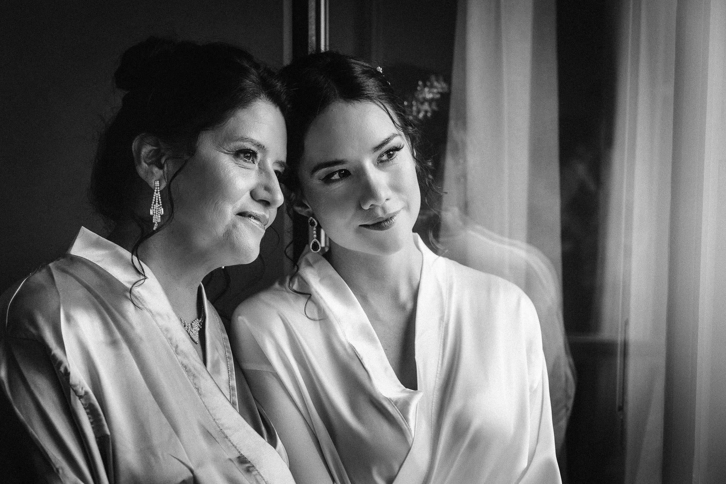 A bride and her mother standing together, both wearing robes, and gazing out of a window with expressions of warmth and closeness.
