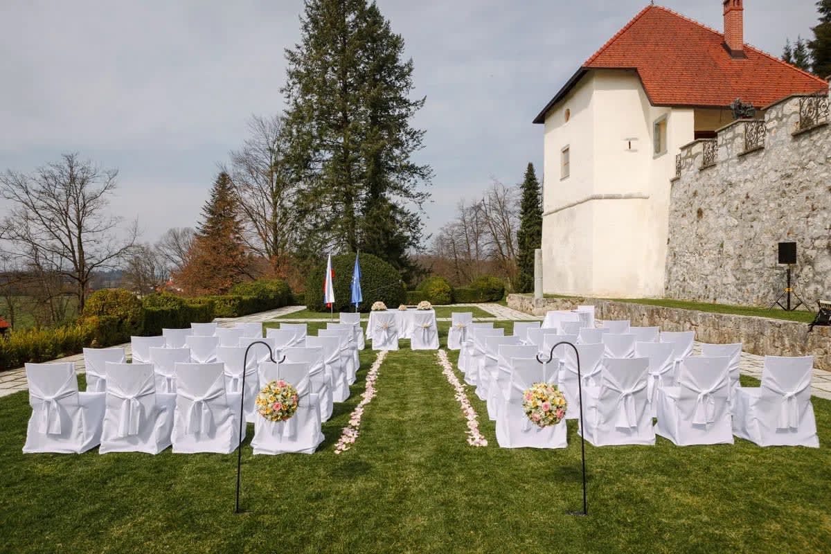 An outdoor wedding ceremony setup with white chairs and floral decorations in the garden of a castle.